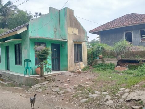 Kondisi luar perpustakaan: Fasad bangunan hijau-krem retak parah. Halaman depan berantakan dengan puing dan pagar tanah di sisi kanan jebol.
